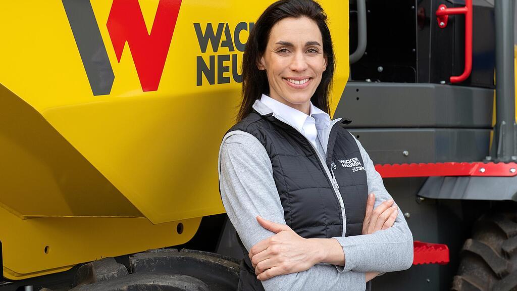 Une collaboratrice Wacker Neuson souriante et les bras croisés devant un dumper sur pneus de Wacker Neuson.