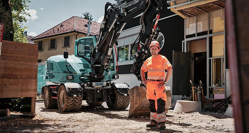 Un opérateur vêtu d'une tenue de travail haute visibilité se tient à côté d'une pelle sur pneus Wacker Neuson EW100 sur un chantier urbain.