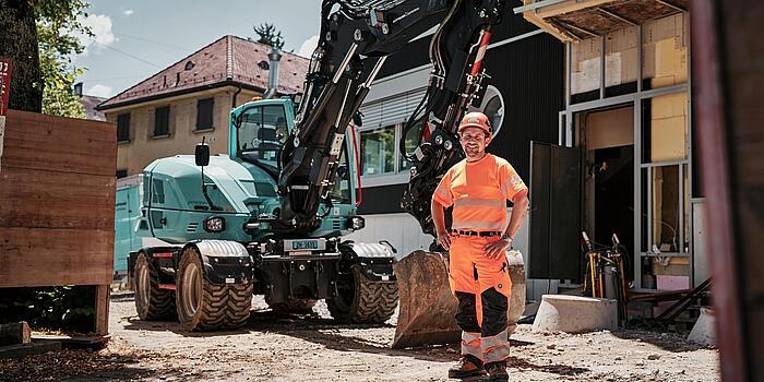 Un opérateur vêtu d'une tenue de travail haute visibilité se tient à côté d'une pelle sur pneus Wacker Neuson EW100 sur un chantier urbain.