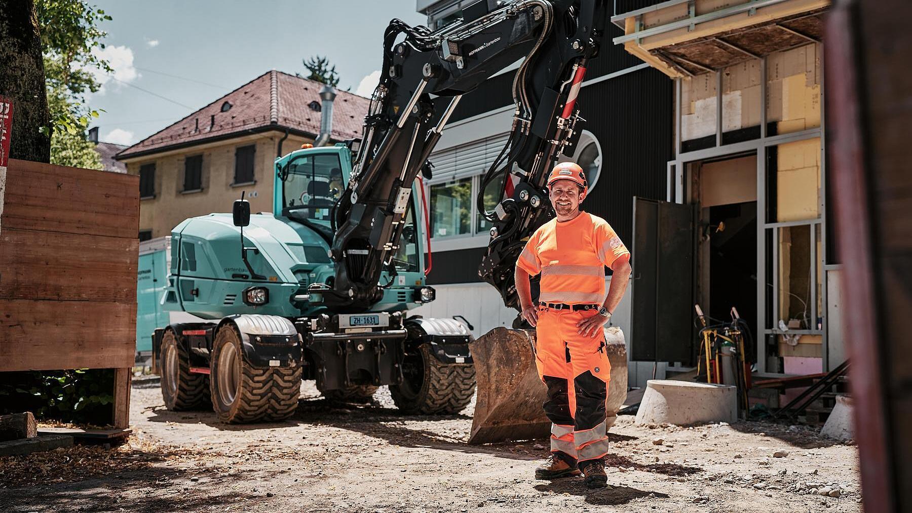 Un opérateur vêtu d'une tenue de travail haute visibilité se tient à côté d'une pelle sur pneus Wacker Neuson EW100 sur un chantier urbain.