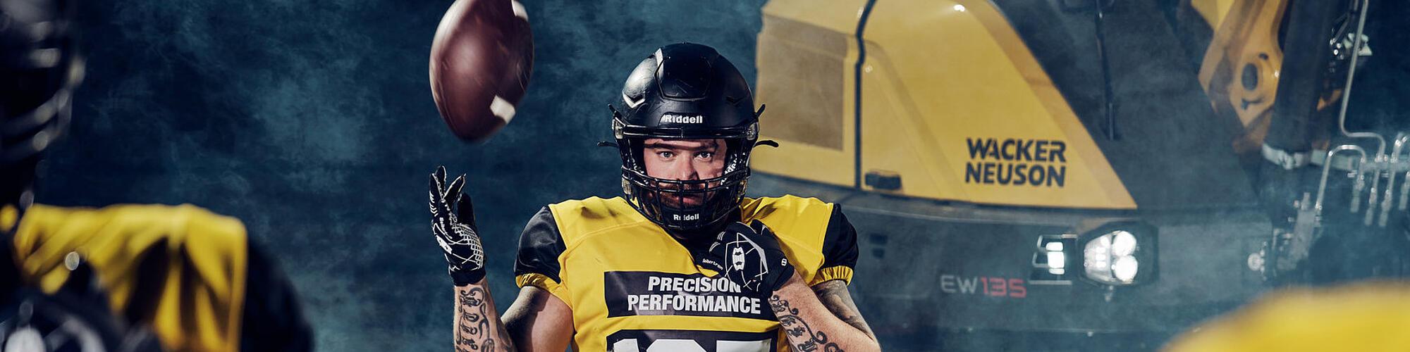 American football player in a “Precision Performance” jersey catching a football in front of a Wacker Neuson EW135 wheeled excavator.