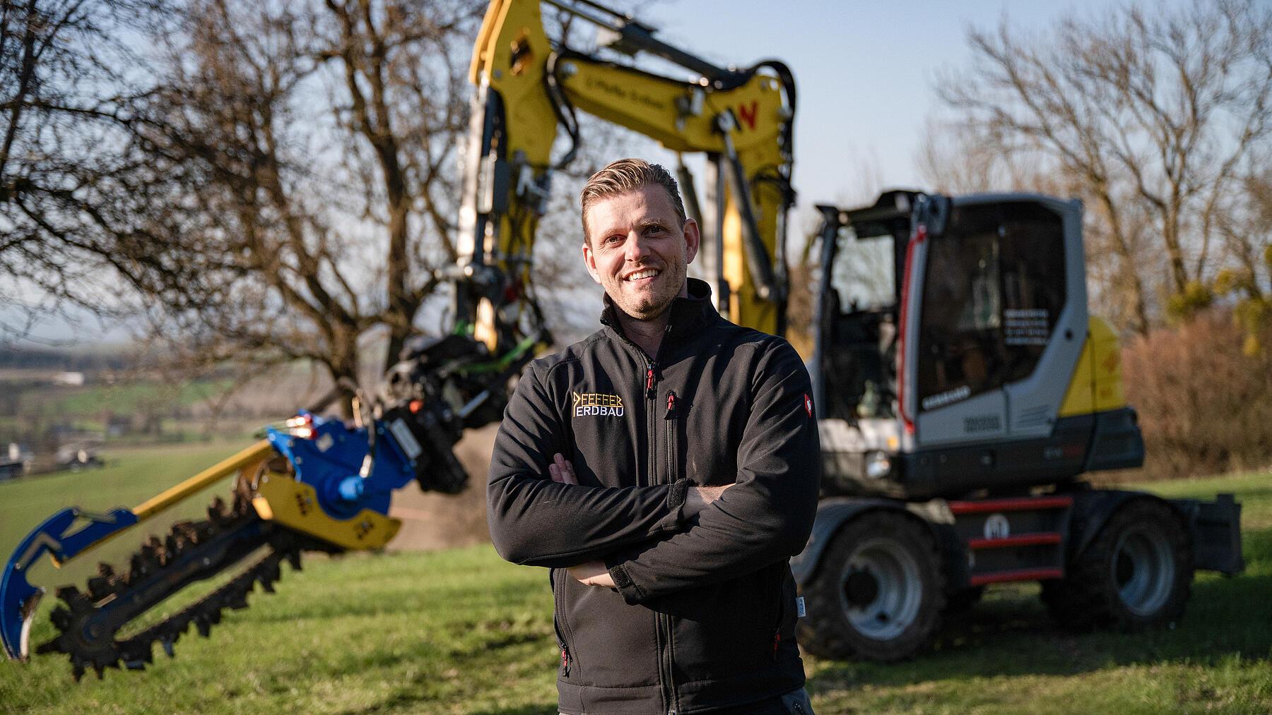 Un entrepreneur se tient devant une pelle sur roues Wacker Neuson EW100 équipée d'un accessoire de creusement de tranchées, sur un chantier en milieu rural.