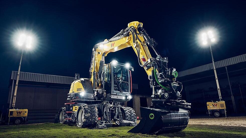 Pelle sur pneus Wacker Neuson EW135 équipée d'un rotateur inclinable et d'un godet de nivellement, éclairée par des projecteurs dans un environnement de travail nocturne.c