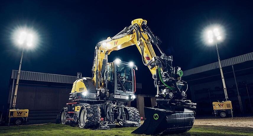 Pelle sur pneus Wacker Neuson EW135 équipée d'un rotateur inclinable et d'un godet de nivellement, éclairée par des projecteurs dans un environnement de travail nocturne.c