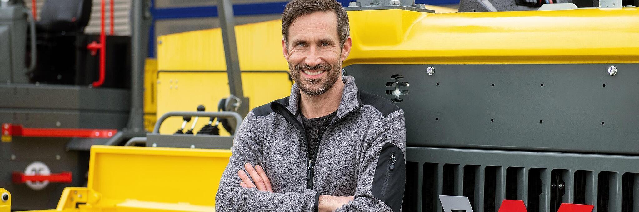 Un collaborateur Wacker Neuson souriant et bras croisés s’appuie sur une machine de construction Wacker Neuson.