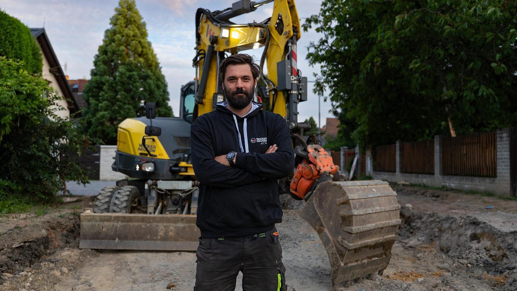 Un professionnel du bâtiment se tenant sur un chantier devant une pelle sur pneus Wacker Neuson EW100.